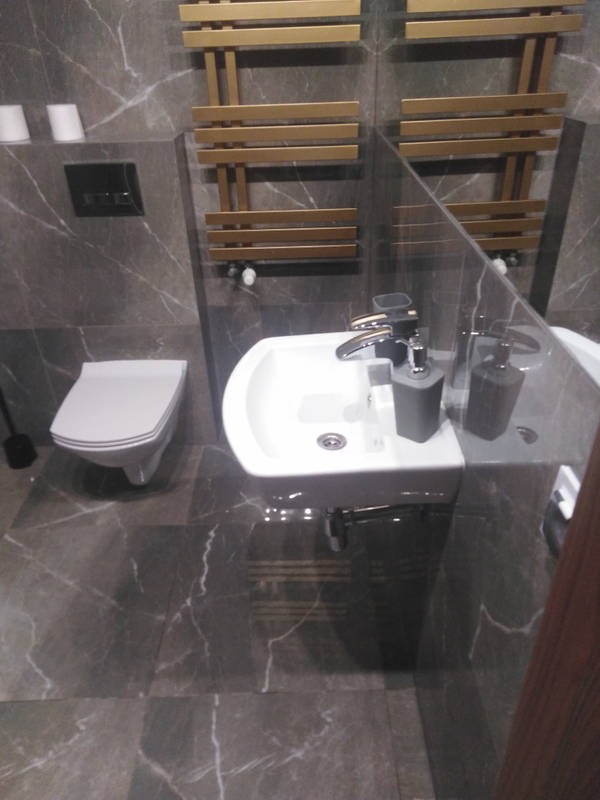 Nowoczesna łazienka z szarymi marmurkowymi płytkami, wisząca toaleta, biała umywalka nablatowa z szarą armaturą i złotym grzejnikiem drabinkowym.