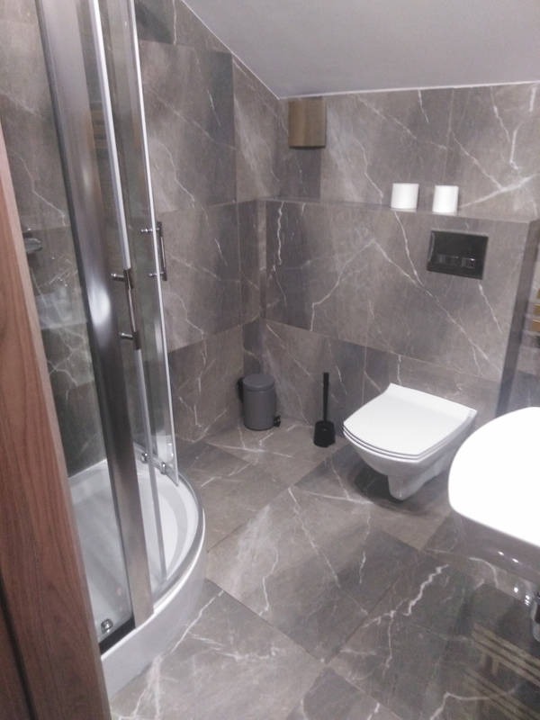 Wykończona łazienka z szarymi płytkami imitującymi marmur na ścianach i podłodze, kabina prysznicowa z zaokrąglonymi drzwiami, wisząca toaleta oraz kosz na śmieci w rogu.