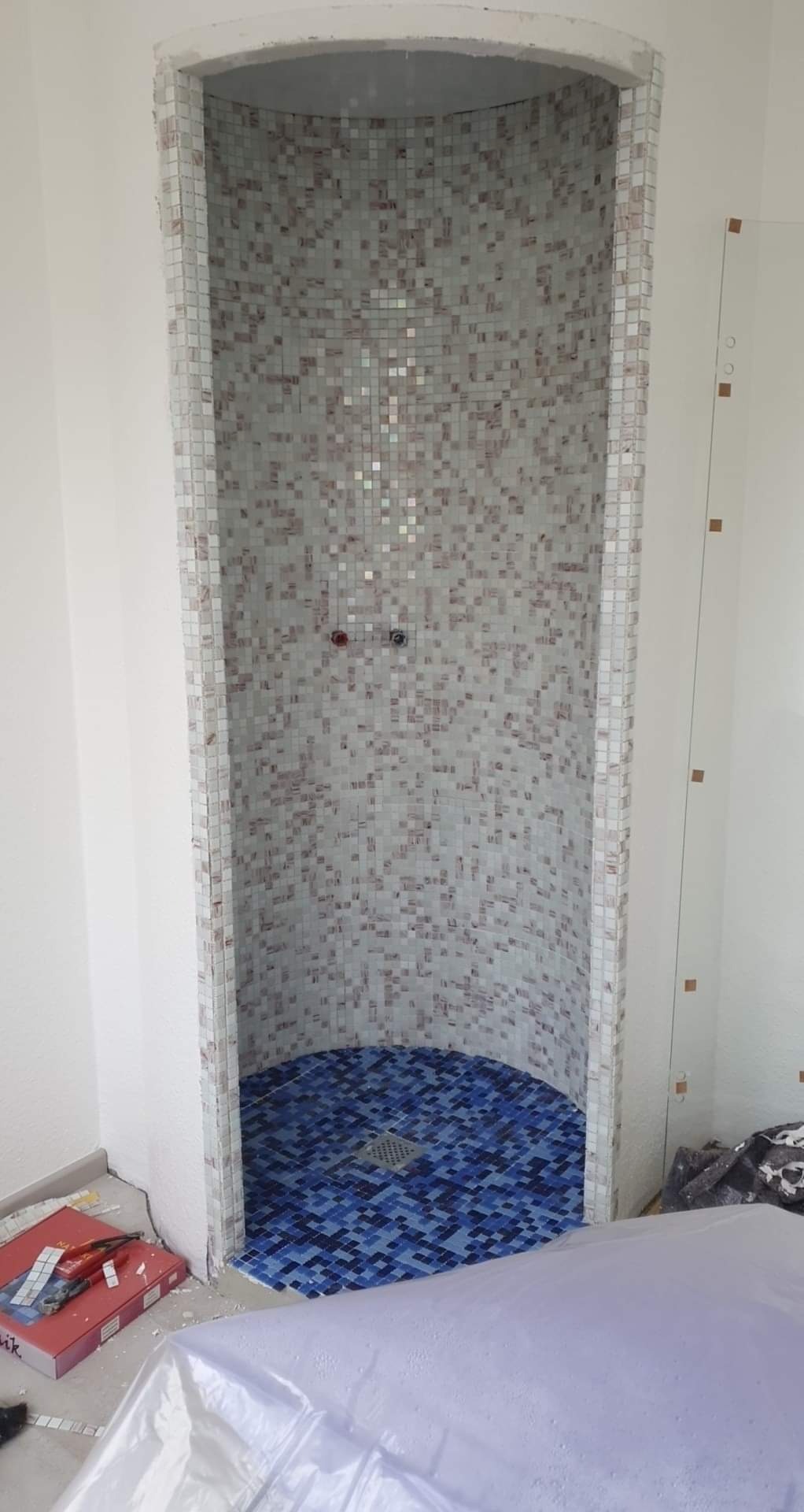 Półokrągła wnęka prysznicowa w trakcie wykańczania mozaiką w odcieniach bieli i beżu, z niebieską mozaiką na podłodze i widocznymi narzędziami glazurniczymi obok.