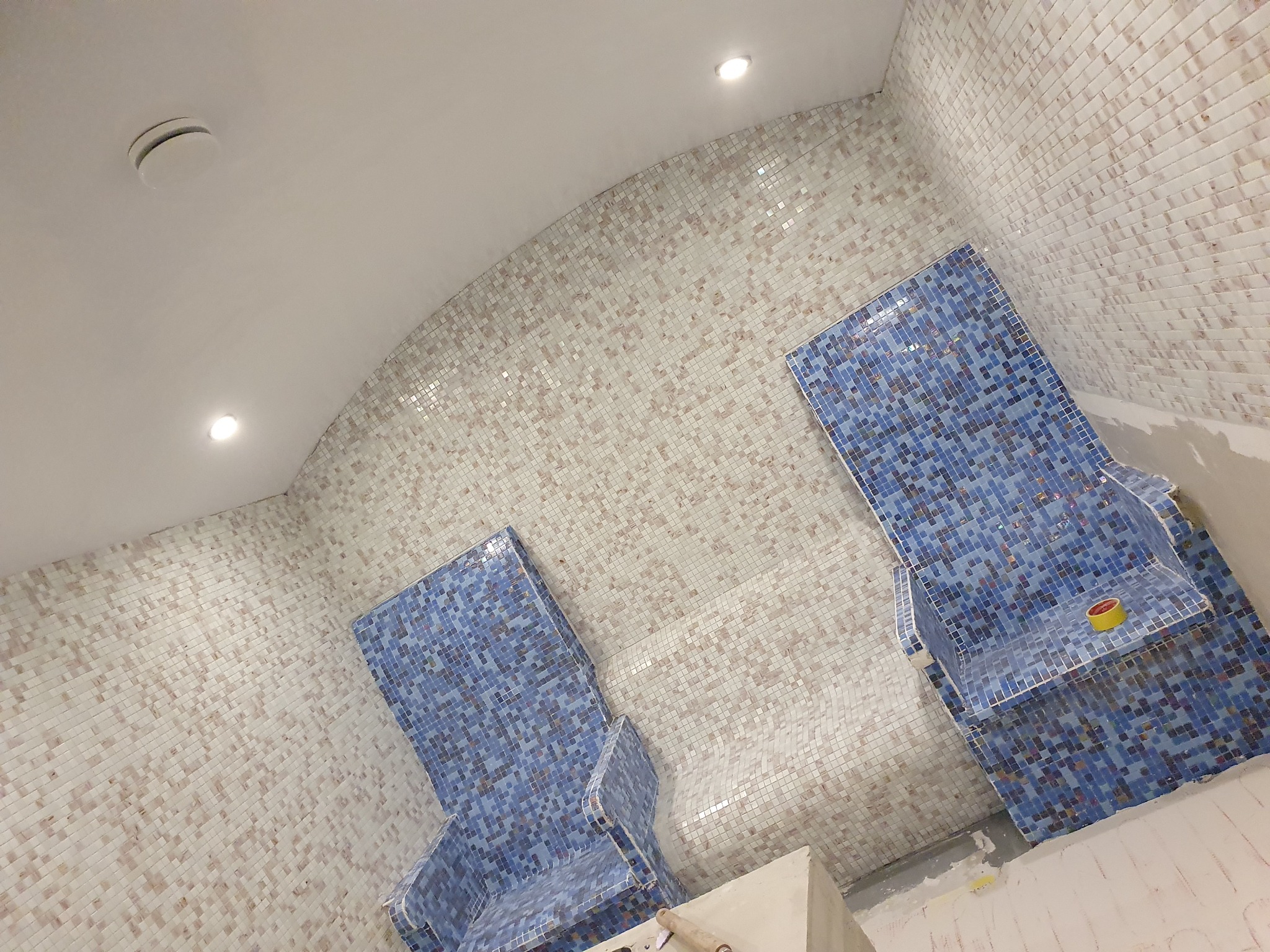 Mozaika w odcieniach bieli i beżu na ścianach łazienki w trakcie prac wykończeniowych, z dwoma wnękami/siedziskami wyłożonymi niebieską mozaiką, taśma malarska na jednym z siedzisk.