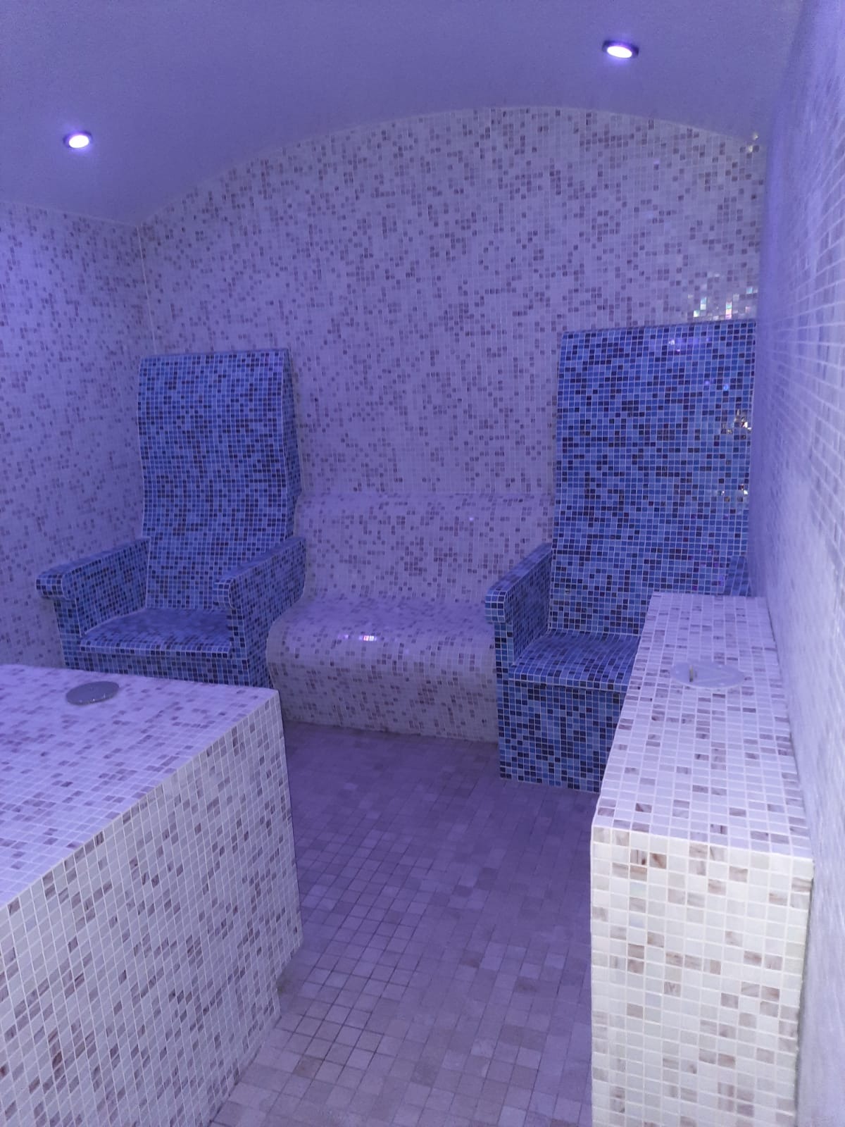 Mozaika w odcieniach błękitu i beżu pokrywająca ściany, podłogę, siedziska i dwa wysokie krzesła w saunie parowej oświetlonej fioletowym światłem.