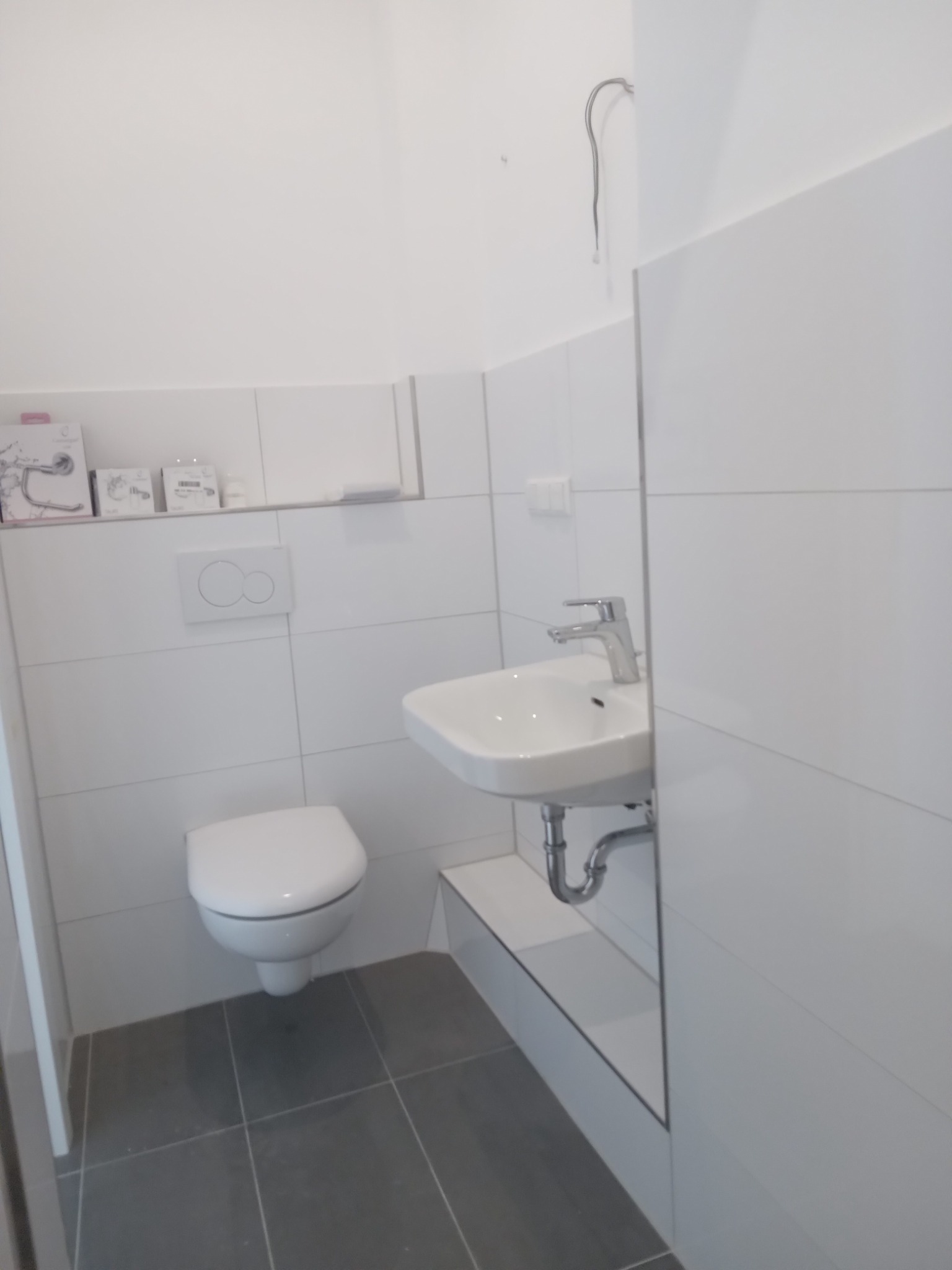 Nowoczesna, minimalistyczna łazienka z białymi płytkami i szarą podłogą. Umywalka nablatowa z chromowaną baterią, wisząca toaleta i akcesoria łazienkowe na półce.