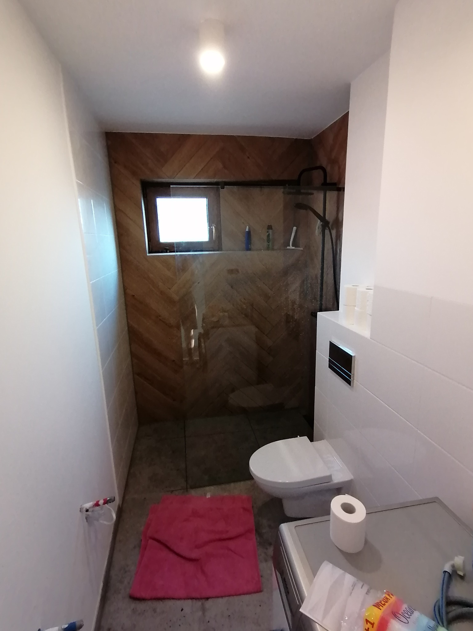 Nowoczesna łazienka z kabiną prysznicową typu walk-in, wykończona drewnem ułożonym w jodełkę na ścianie prysznica, biała ceramika sanitarna, widoczny fragment pralki z rolką papieru toaletowego...