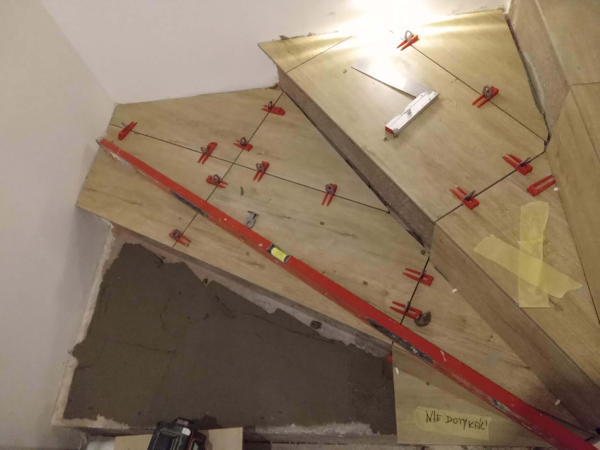Montaż płytek na schodach z użyciem systemu poziomowania płytek (czerwone klipsy i kliny), poziomica w kolorze czerwonym, widoczna zaprawa murarska i kartka z napisem 'Nie dotykać!'