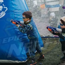 Dwaj chłopcy w okularach ochronnych celują z pistoletów na kulki podczas zabawy w plenerze, widoczny dmuchany obiekt z logo Epic Nerf i porozrzucane naboje.