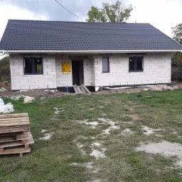 Surowy dom murowany w trakcie budowy z ciemną dachówką, widoczne bloczki, czarne ramy okienne, palety na trawie i żółta tablica informacyjna przy wejściu.