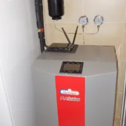 Szara jednostka grzewcza z czerwonym panelem, czarnym zbiornikiem wyrównawczym i przewodami w tle, router Wi-Fi leżący na górze urządzenia.