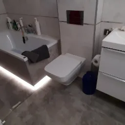 Nowoczesna łazienka z podświetlaną wanną, umywalką z szafką i toaletą w odcieniach szarości i bieli, uzupełniona o akcesoria łazienkowe.