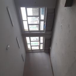 Łukaszbud - Wnętrze remontowanego pomieszczenia z dwoma oknami, grzejnikami i widocznymi śladami po gładzi na ścianach i podłodze, oświetlone listwami LED.