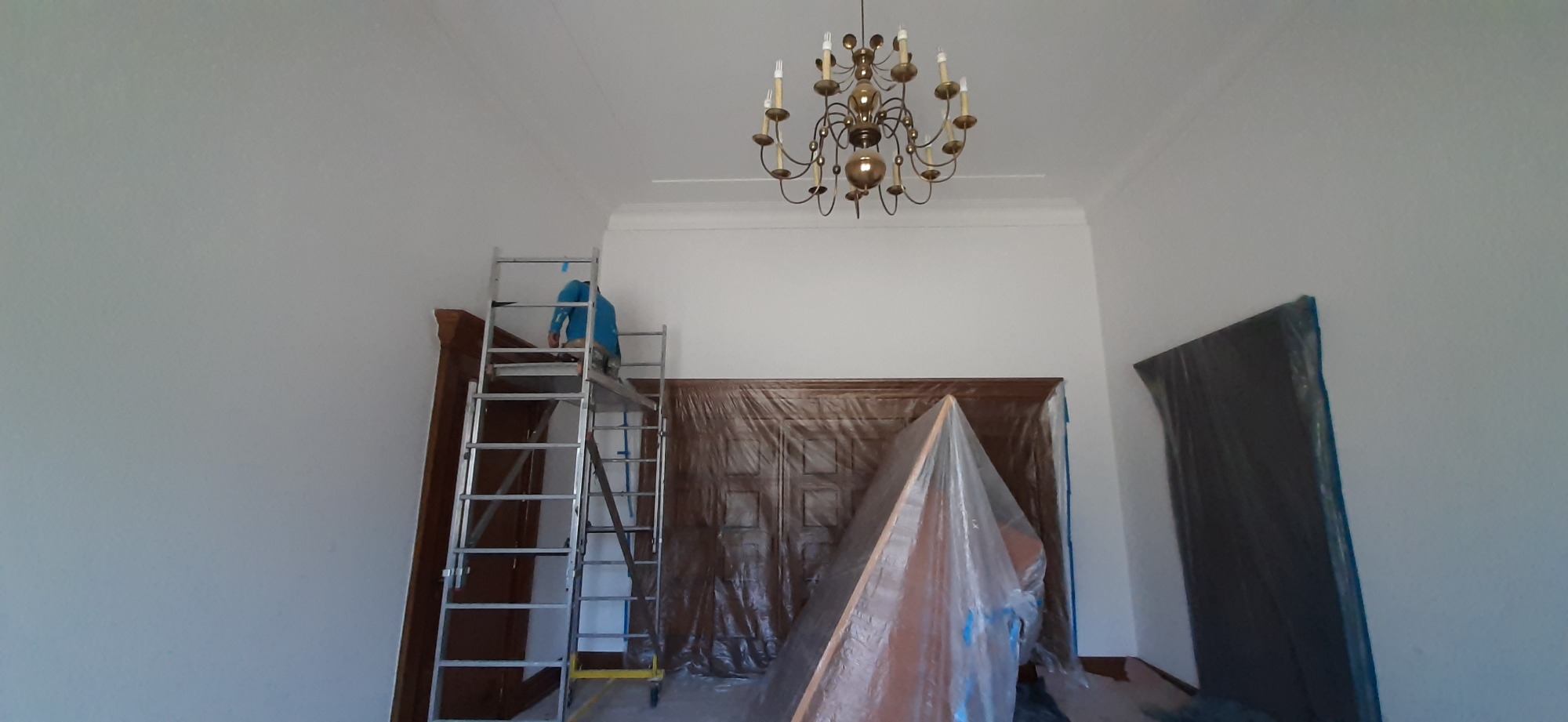 Mężczyzna na rusztowaniu maluje ścianę w pomieszczeniu z żyrandolem, meble i drzwi zabezpieczone folią malarską.