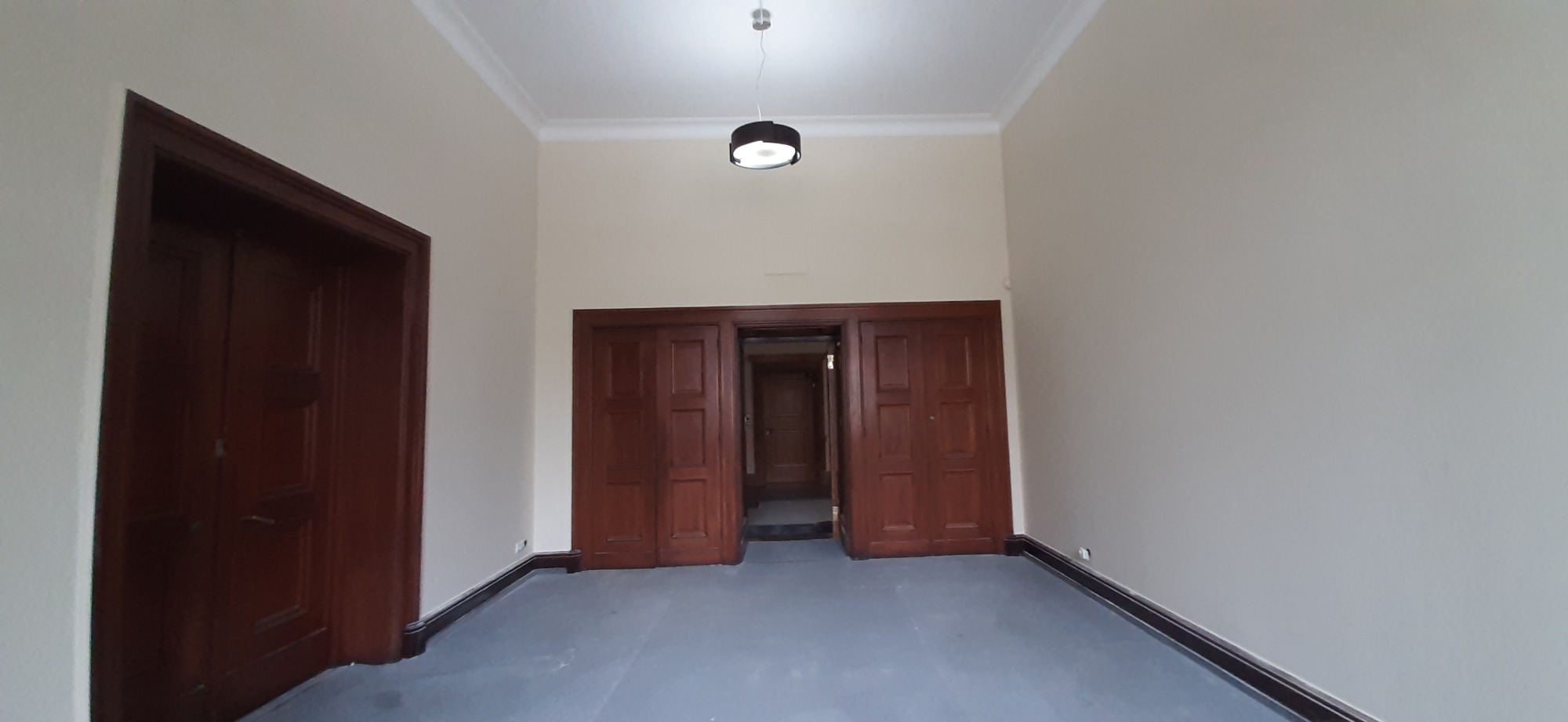 Wnętrze pokoju z odświeżonymi ścianami w odcieniach beżu, ciemnobrązowymi drewnianymi drzwiami i listwami przypodłogowymi oraz nowoczesną lampą sufitową.