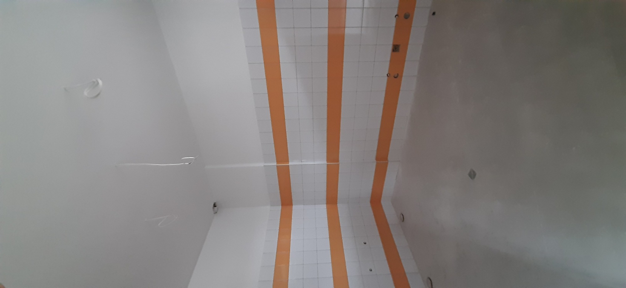 Wnętrze pomieszczenia z przygotowanymi ścianami do dalszych prac wykończeniowych, widoczne ślady po przewodach elektrycznych na suficie i częściowo wykończona ściana z pomarańczowymi pasami.