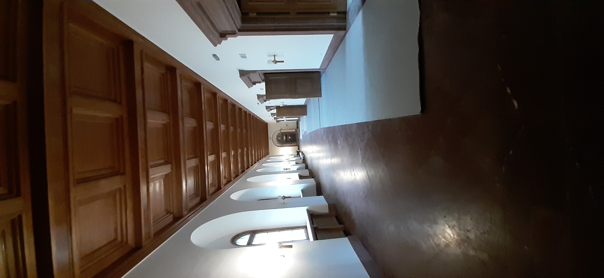 Długi korytarz z drewnianą boazerią, białymi ścianami z łukami i oknami, oraz polerowaną podłogą, perspektywa z jednego końca.