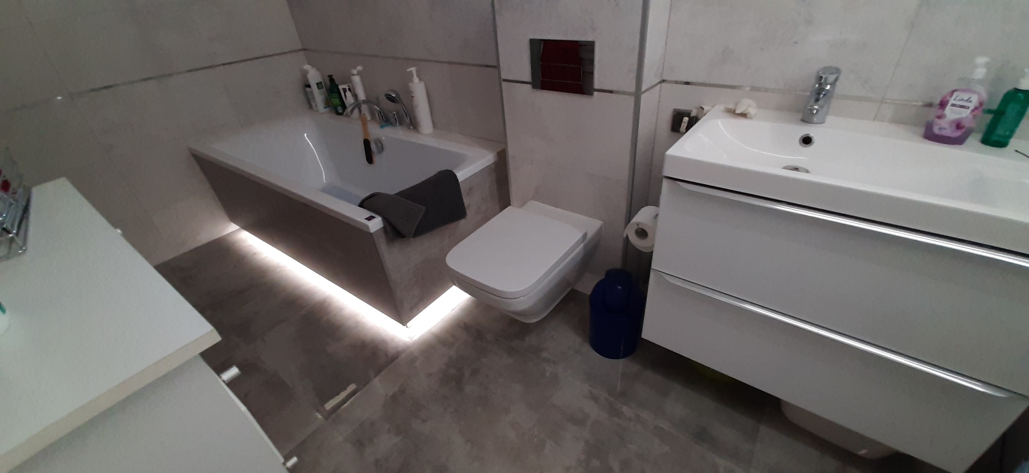 Nowoczesna łazienka z podświetlaną wanną, umywalką z szafką i toaletą w odcieniach szarości i bieli, uzupełniona o akcesoria łazienkowe.