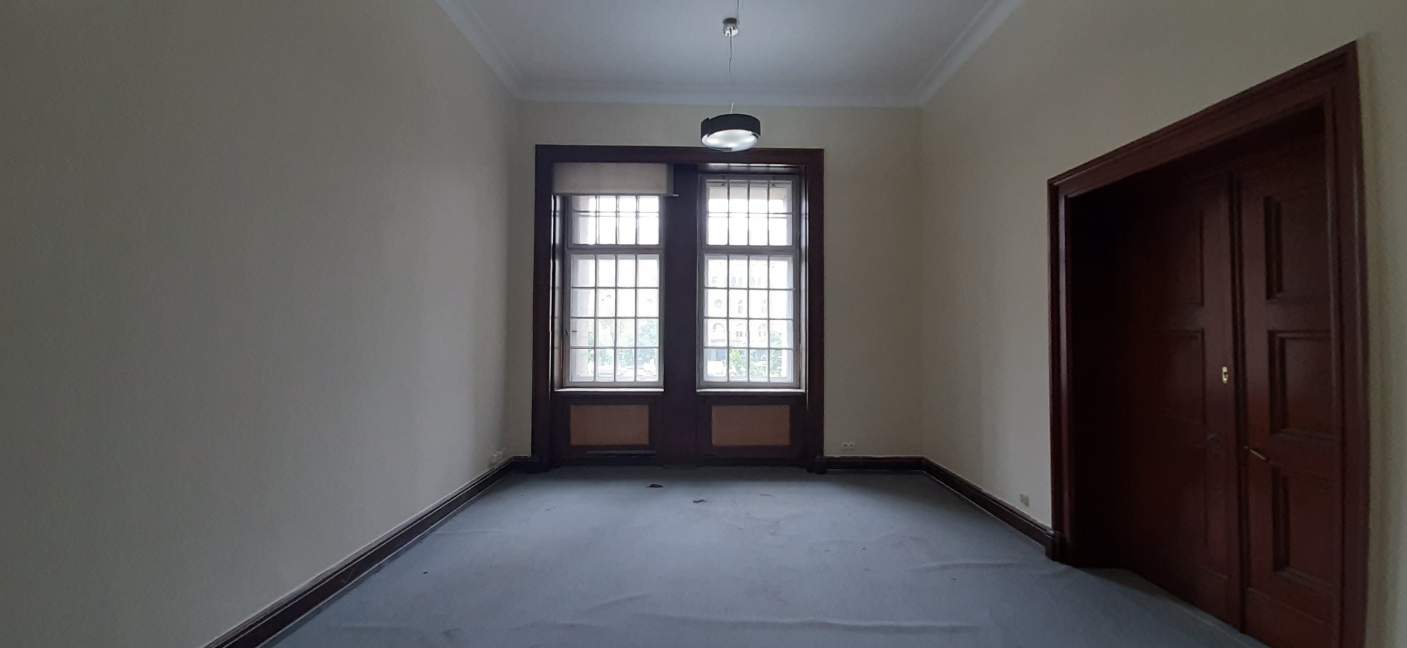 Puste, jasne pomieszczenie z kremowymi ścianami, ciemnymi listwami przypodłogowymi, podwójnymi drzwiami balkonowymi z roletą i ciemnymi ramami, oraz ciemnymi drzwiami po prawej stronie, oświetlone...