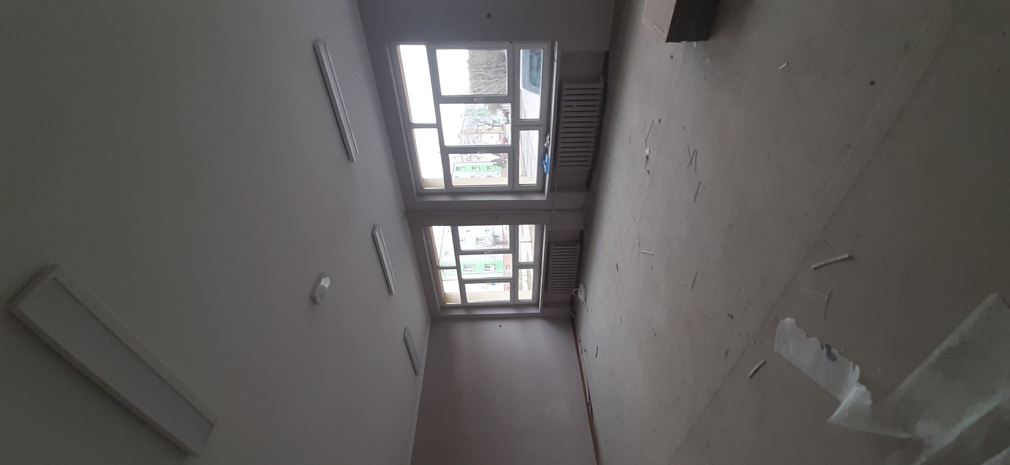 Wnętrze remontowanego pomieszczenia z dwoma oknami, grzejnikami i widocznymi śladami po gładzi na ścianach i podłodze, oświetlone listwami LED.