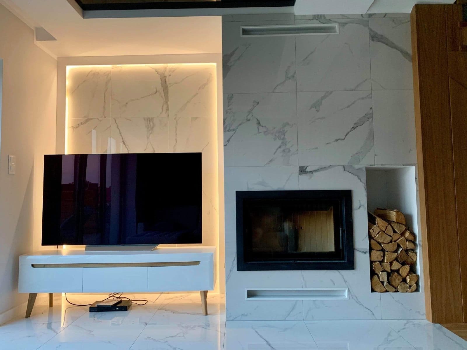 Nowoczesny salon z marmurowymi płytkami na ścianie, kominkiem z czarną obudową i ułożonym drewnem, telewizorem na białej szafce RTV z drewnianymi nóżkami, oraz podświetleniem LED za panelem ściennym.