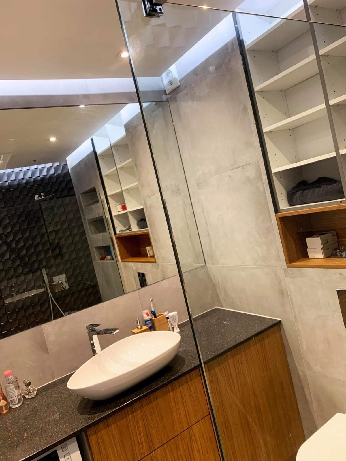 Nowoczesna łazienka z umywalką nablatową o owalnym kształcie, baterią chromowaną i blatem z ciemnego kamienia, na tle szarych płytek imitujących beton i czarnej ściany z geometrycznym wzorem.