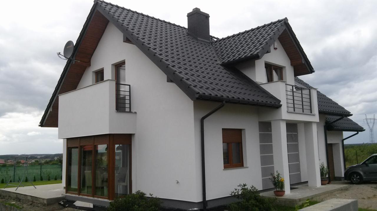 Nowoczesny, dwupiętrowy dom jednorodzinny z grafitową dachówką, białymi ścianami i drewnianymi oknami, widziany z zewnątrz w pochmurny dzień.