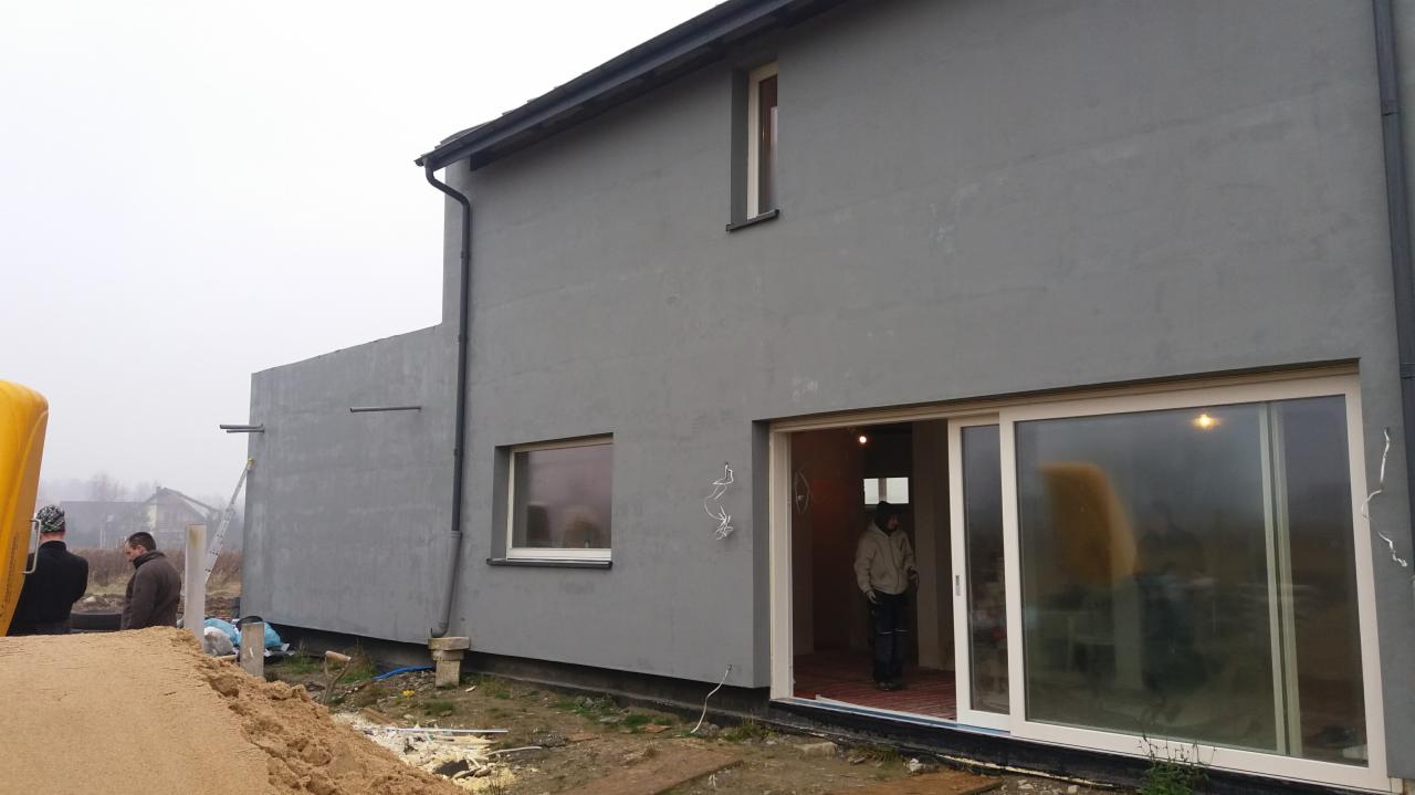 Szary budynek w trakcie budowy z widocznymi oknami, drzwiami przesuwnymi, izolacją przy fundamentach i dwoma mężczyznami na zewnątrz, z żółtym zbiornikiem w tle.