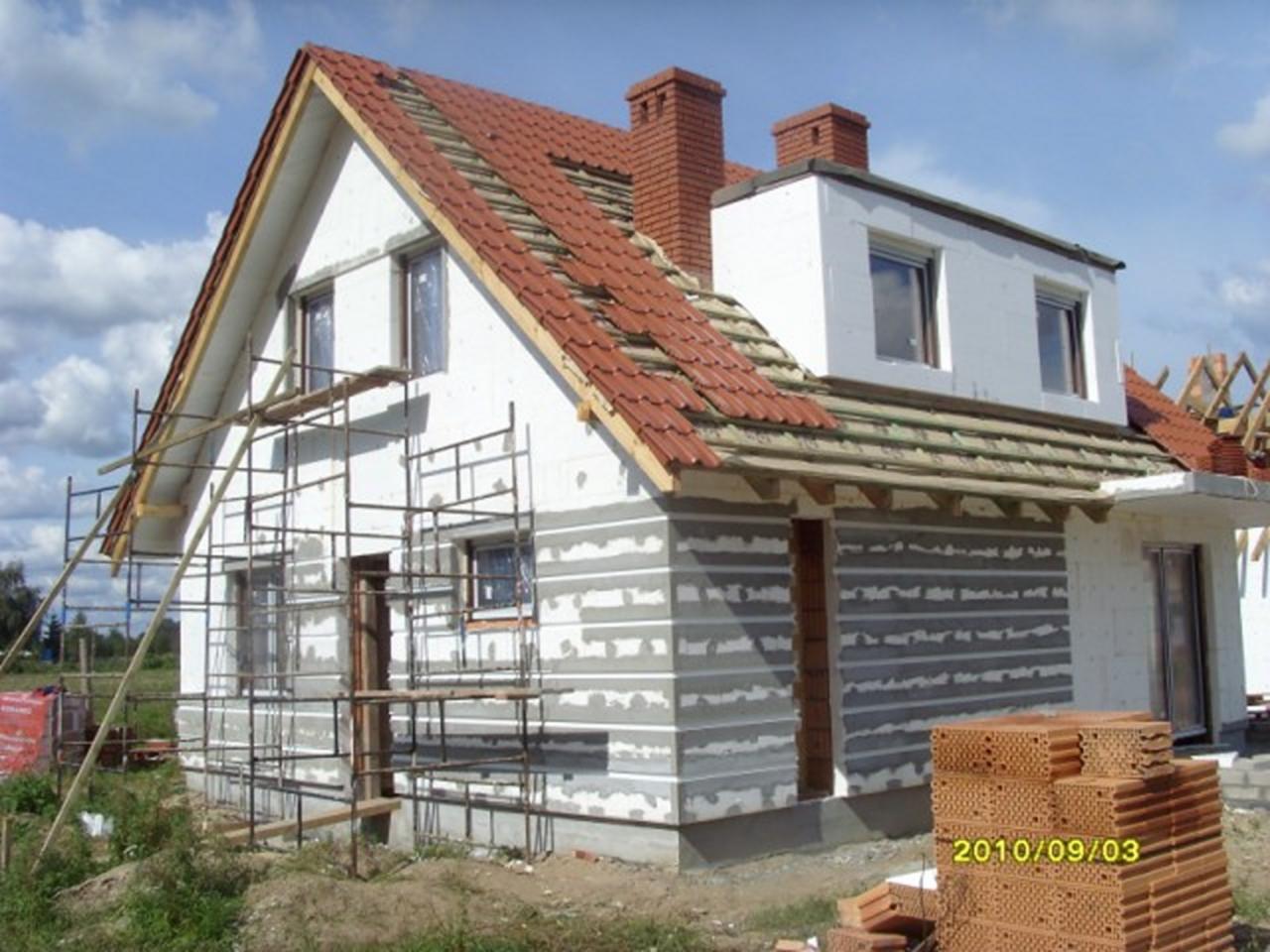 Budowa domu jednorodzinnego w trakcie ocieplania ścian zewnętrznych, widoczne rusztowania, częściowo położona dachówka i ułożone bloczki ceramiczne.