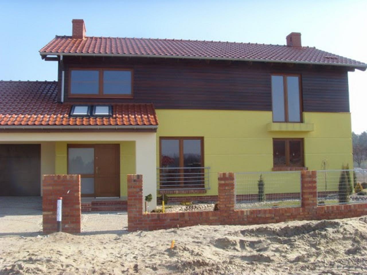 Dwukondygnacyjny dom w Szczecinie z żółtą elewacją i brązowym drewnianym wykończeniem górnej części, czerwonym dachem z dachówki i ceglanym ogrodzeniem w trakcie budowy.