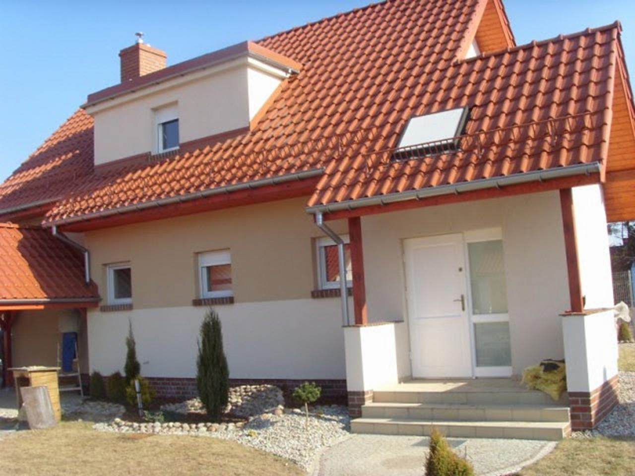 Dom jednorodzinny z czerwonym dachem z dachówki, jasną elewacją w dwóch odcieniach beżu i białym wejściem z kolumnami, widziany z perspektywy ogrodu.