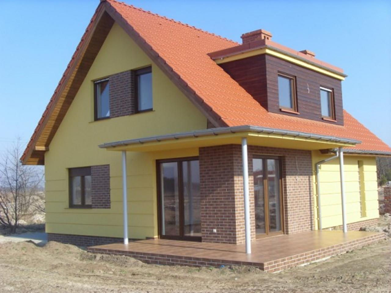 Żółty dom jednorodzinny z czerwoną dachówką, brązowymi oknami i drzwiami, oraz częściowo wykończoną elewacją z cegły klinkierowej, widoczny w słoneczny dzień.