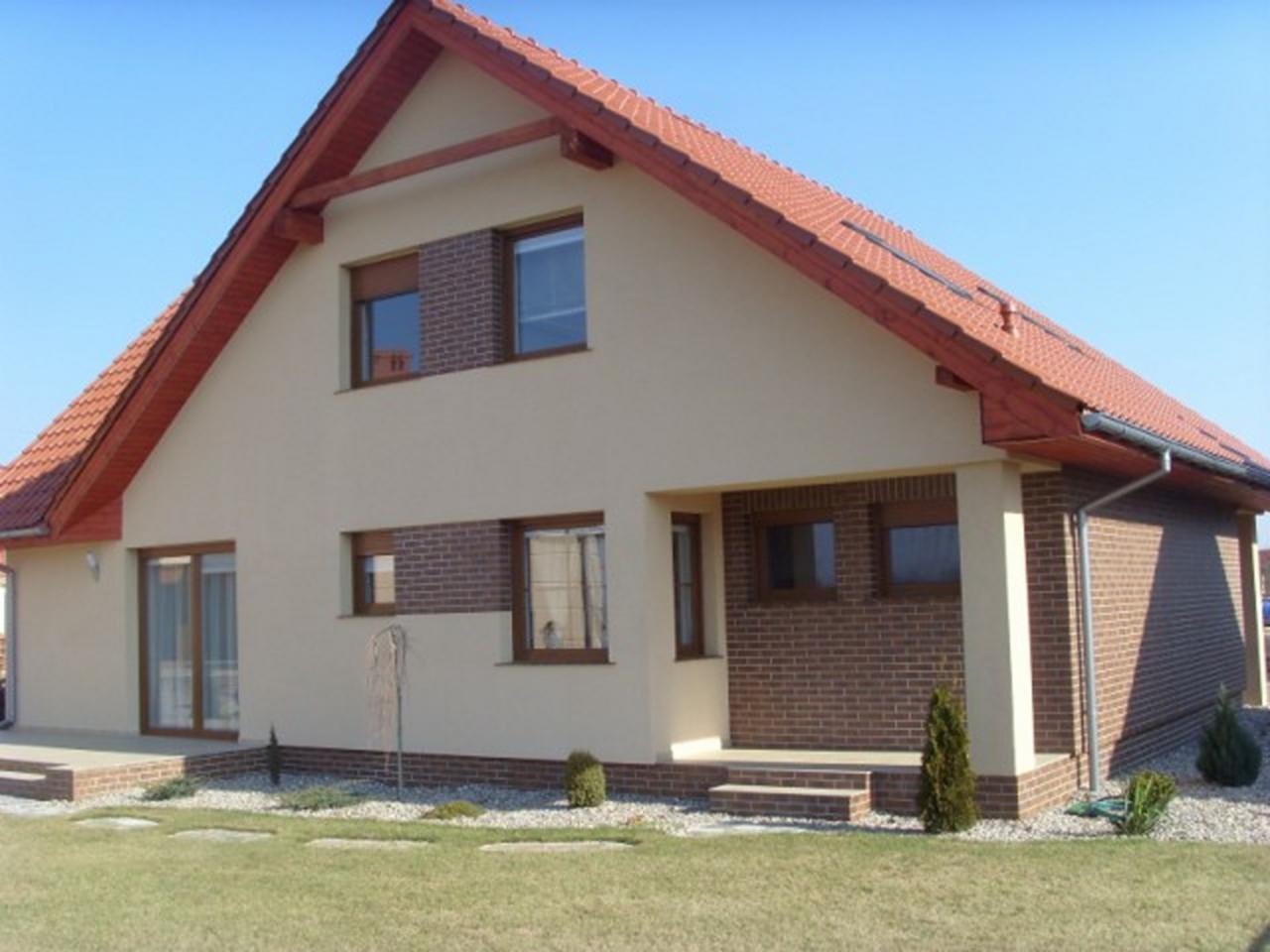 Nowoczesny dom jednorodzinny z beżową elewacją i fragmentami muru z cegły klinkierowej, czerwona dachówka, jasne okna, trawnik z ozdobnymi kamieniami i niskimi krzewami.