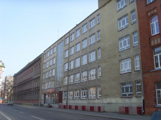 Budynek szkoły z widocznymi oknami i elewacją w odcieniach szarości i beżu, sąsiadujący z budynkiem z czerwonej cegły, sfotografowany z poziomu ulicy w słoneczny dzień.