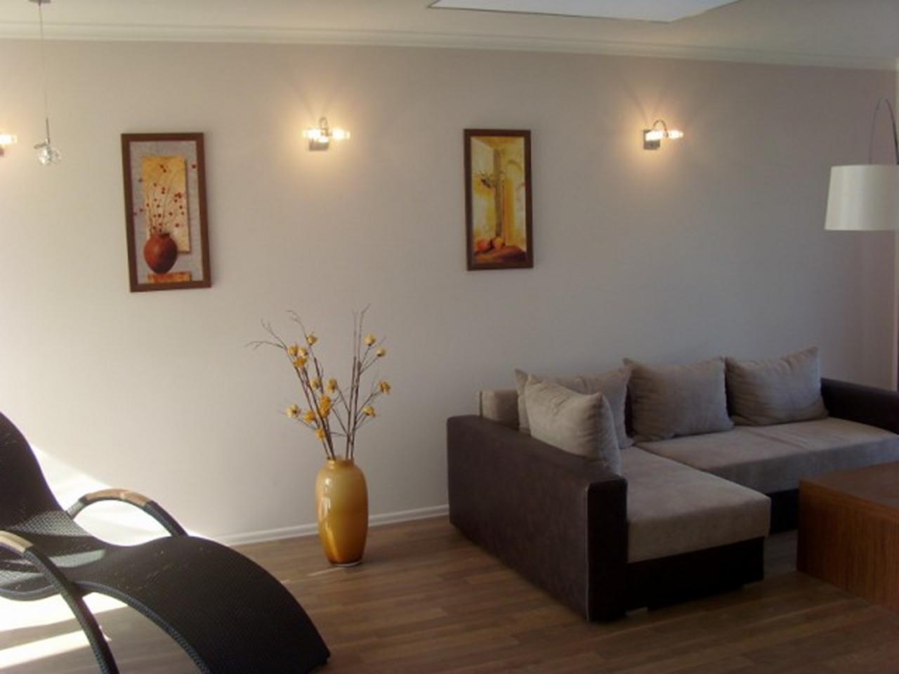 Jasne wnętrze salonu z drewnianą podłogą, szarym narożnikiem, czarnym leżakiem i dekoracyjnym wazonem z żółtymi kwiatami. Na ścianie wiszą obrazy podświetlone kinkietami.