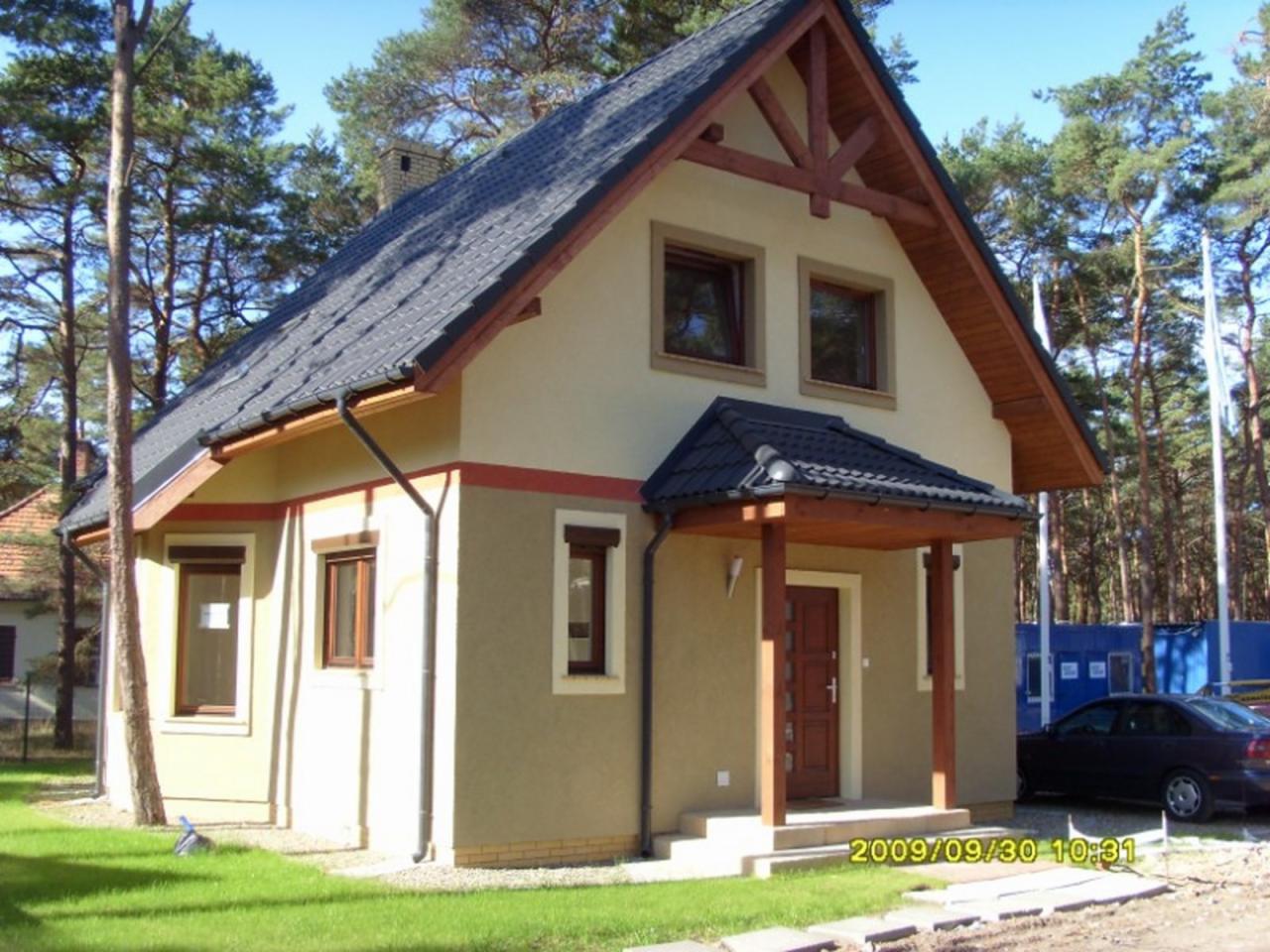 Nowo wybudowany, dwukondygnacyjny dom jednorodzinny z ciemnoszarą dachówką, drewnianą konstrukcją szczytową i zadaszonym wejściem, otoczony lasem sosnowym w słoneczny dzień.