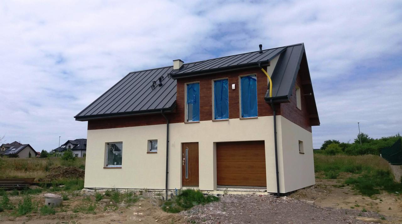 Nowo wybudowany dom jednorodzinny z ciemną blachodachówką, ocieplony styropianem i z częściowo nałożonym tynkiem w kolorze beżowym, okna zabezpieczone folią, zamontowana brama garażowa.
