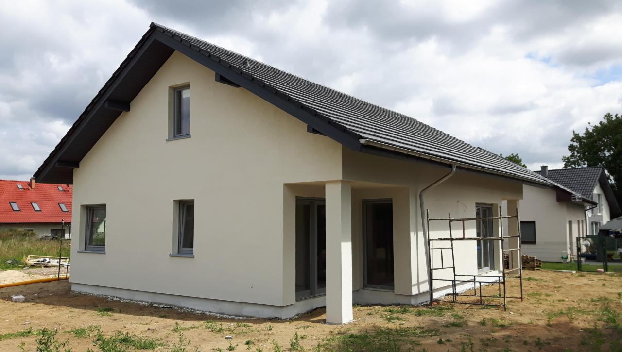 Nowo otynkowany dom jednorodzinny z szarą dachówką i widocznymi rusztowaniami przy elewacji bocznej, ukazujący postęp prac budowlanych.