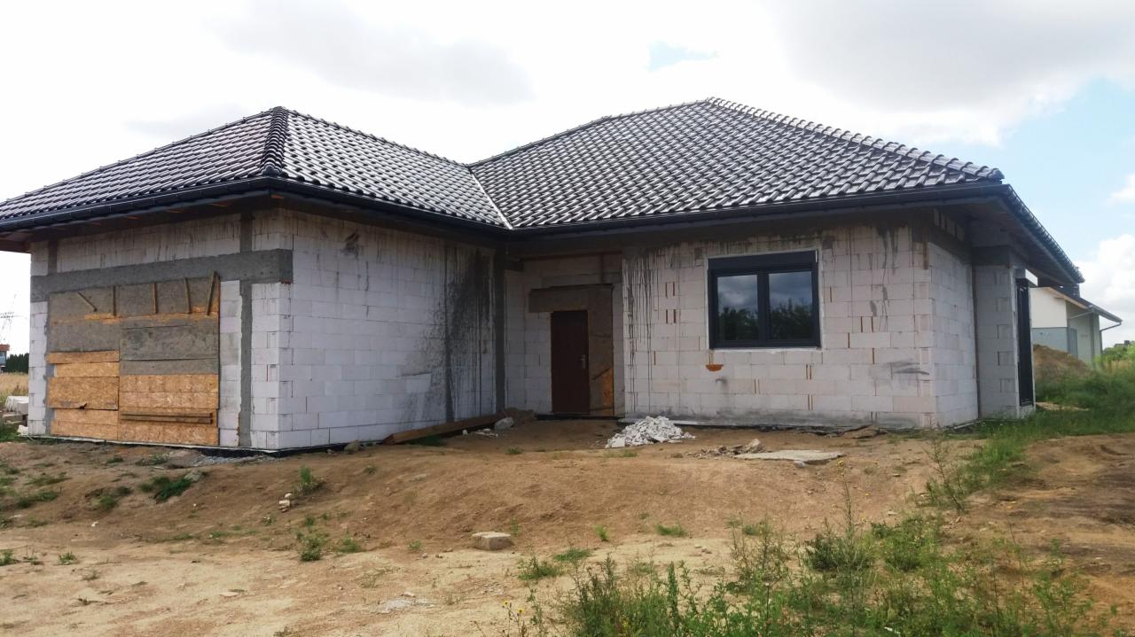 Niedokończony budynek jednorodzinny z ciemną dachówką, widoczne surowe mury z betonu komórkowego, czarne okno i tymczasowe zabezpieczenie otworu garażowego z płyt OSB.