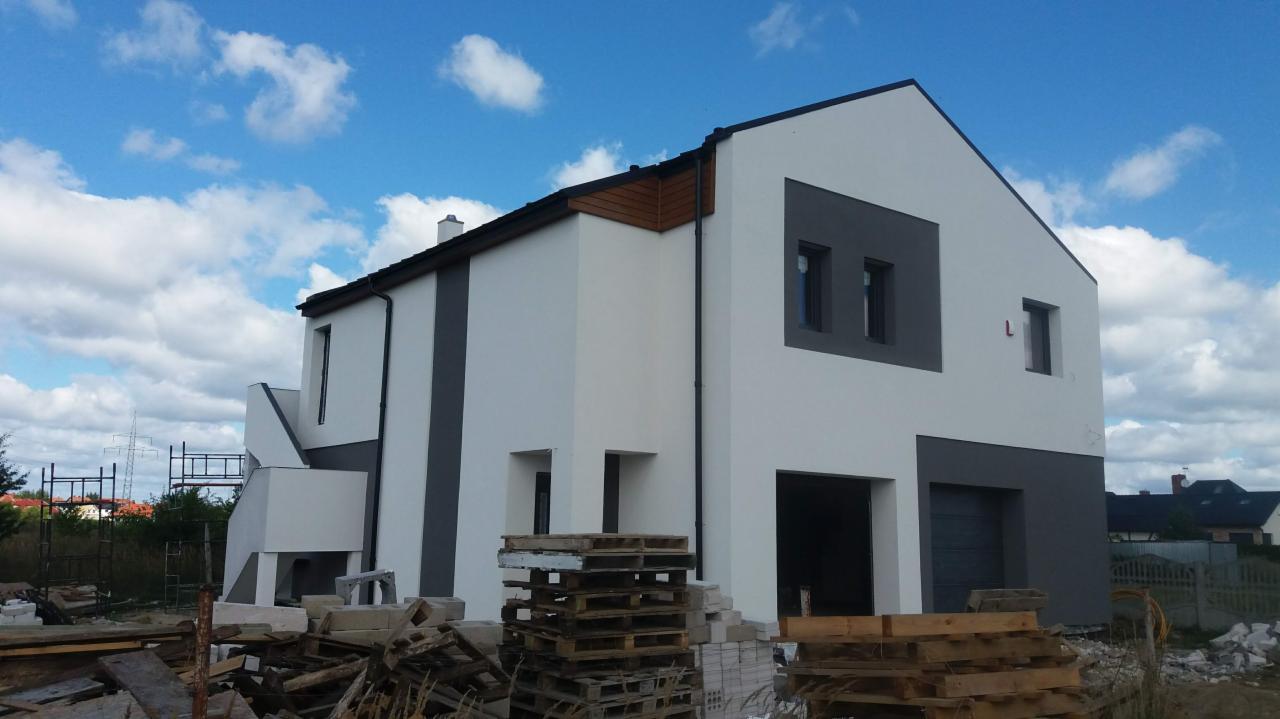 Nowoczesny dom w trakcie wykończenia zewnętrznego z widocznymi paletami i materiałami budowlanymi na pierwszym planie oraz rusztowaniem z boku budynku.