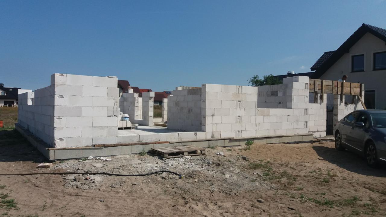 Wznoszenie ścian parteru domu jednorodzinnego z białych bloczków betonowych, widoczne zbrojenie i szalunki nad otworami okiennymi, na pierwszym planie brudna ziemia i zaparkowany samochód osobowy.