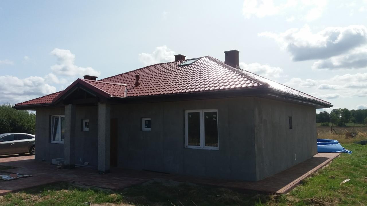 Surowy dom jednorodzinny z czerwoną dachówką i szarym tynkiem zewnętrznym, na etapie budowy, widoczny samochód i basen ogrodowy w tle.