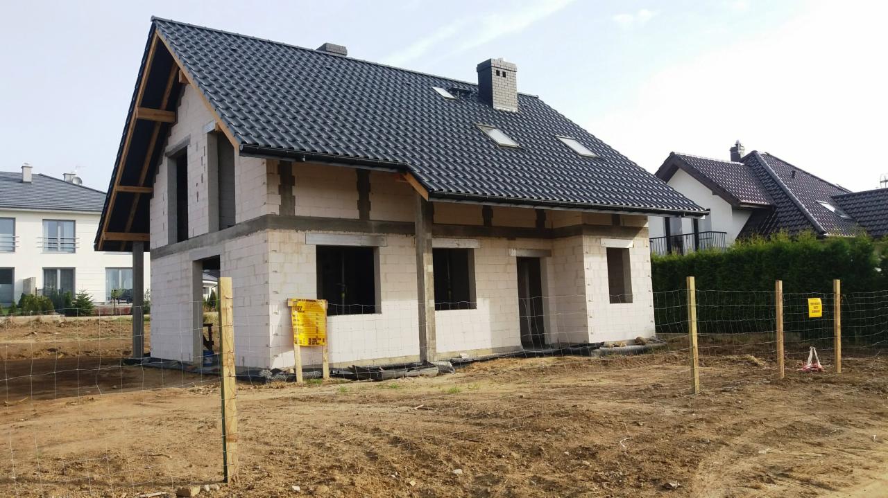 Dom w trakcie budowy z czarną dachówką, widoczne niezabezpieczone otwory okienne i drzwiowe, otoczony siatką ogrodzeniową i żółtymi tablicami informacyjnymi.