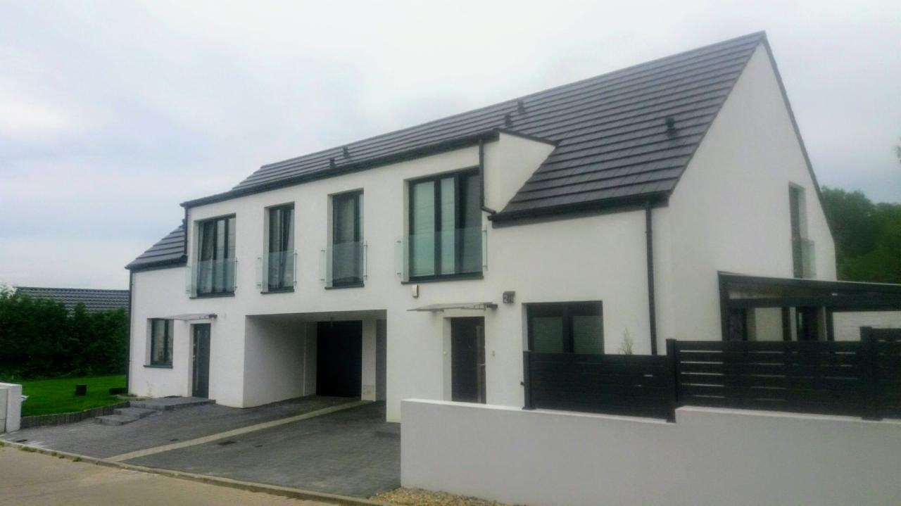 Nowoczesny dwupiętrowy bliźniak z białymi ścianami, grafitowym dachem i czarnymi ramami okiennymi, szklane balustrady balkonowe, widok z zewnątrz w pochmurny dzień.