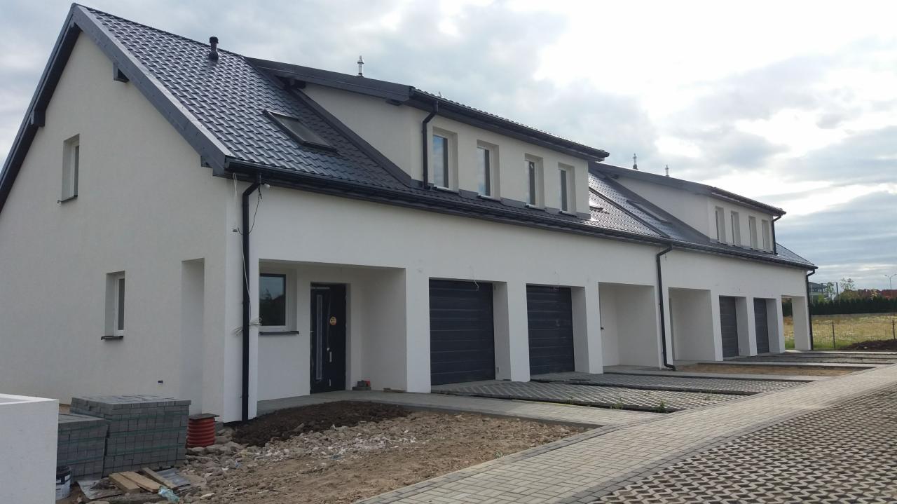 Szeregówka domów jednorodzinnych z czarną dachówką i szarymi bramami garażowymi, widok z boku, z ułożoną kostką brukową i przygotowanym terenem pod zieleń.