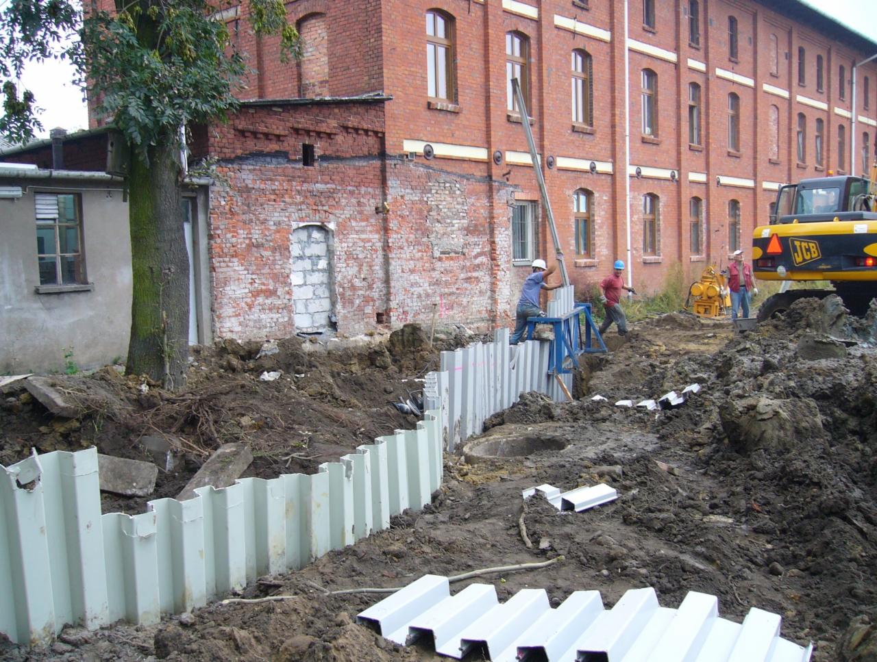 Budowa: pracownicy w kaskach montują stalowe szalunki wykopu na tle ceglanego budynku i koparki JCB.