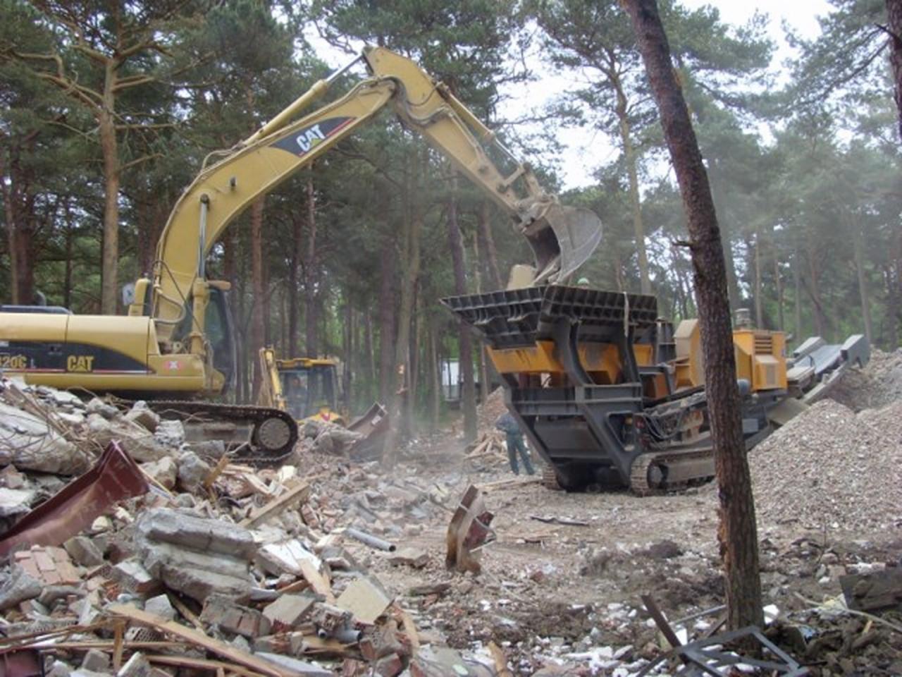 Żółty koparka CAT ładuje gruz do kruszarki na terenie rozbiórki budynku, w tle widoczne drzewa i inne maszyny budowlane.