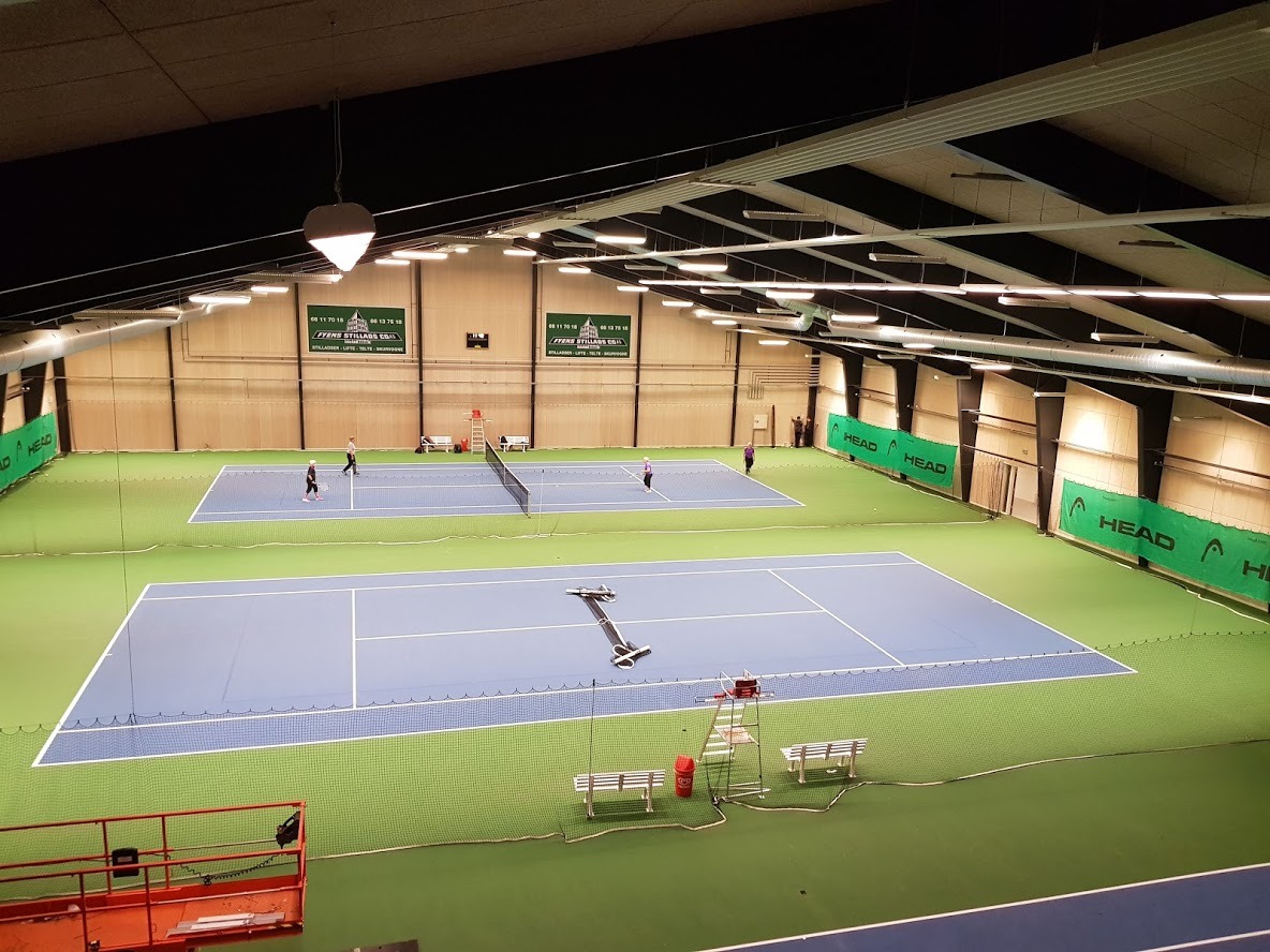 Widok z góry na halę tenisową z dwoma kortami o niebieskiej nawierzchni, zielonym obramowaniem i siatkami, oświetloną lampami sufitowymi, z widocznymi osobami grającymi w tenisa i banerami...