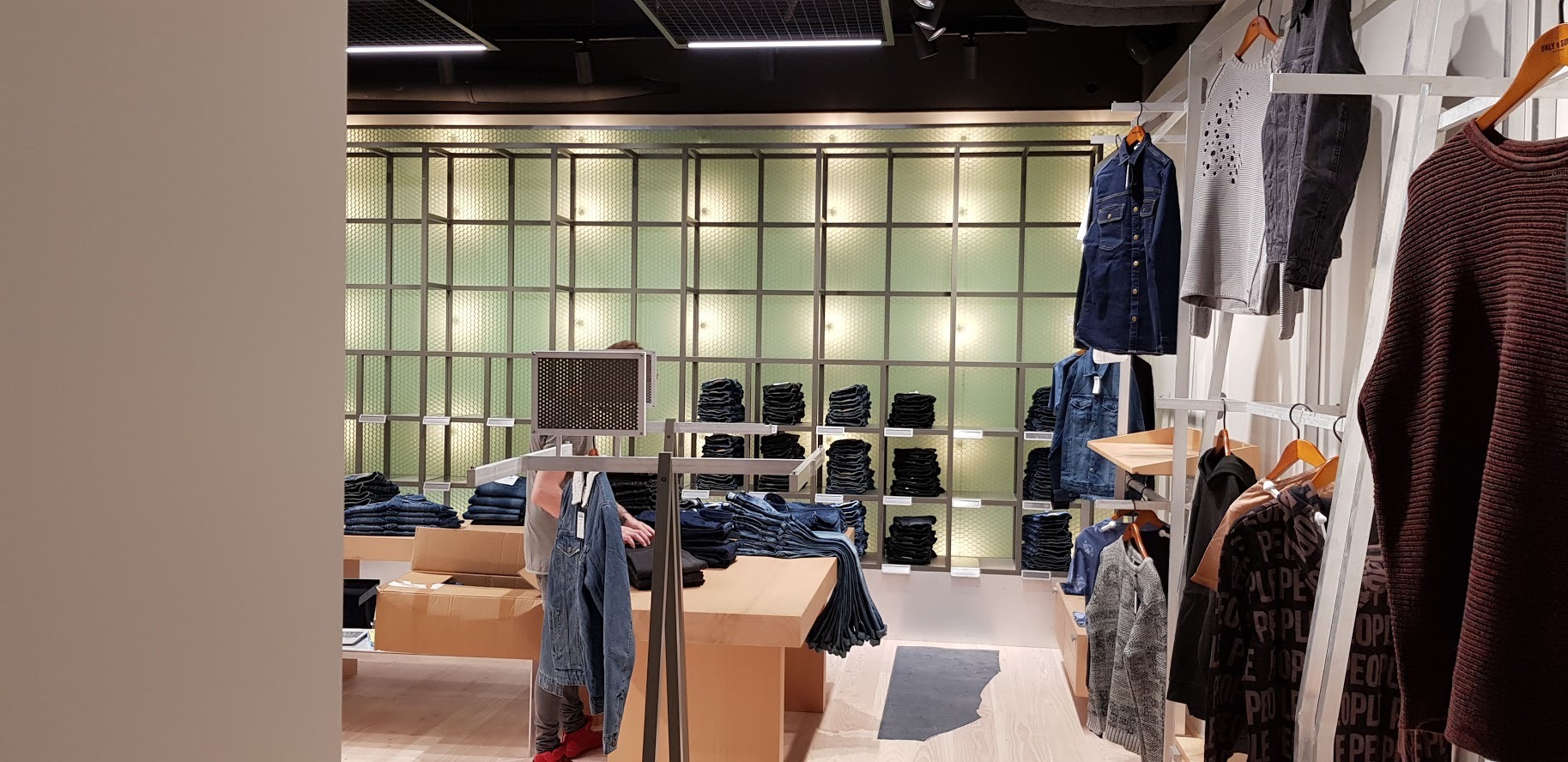 Wnętrze sklepu odzieżowego z półkami i wieszakami prezentującymi poskładane dżinsy i wiszące ubrania, oświetlone punktowo, z pracownikiem układającym towar na stole.