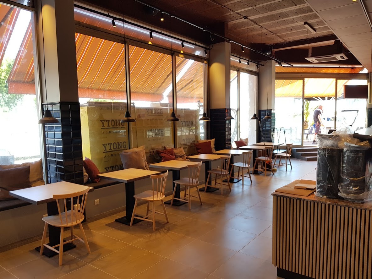 Wnętrze kawiarni z rzędami stolików i krzeseł wzdłuż okien z markizami, widoczne materiały budowlane YTONG, oświetlenie punktowe i wiszące lampy.