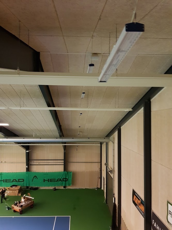 Wnętrze hali sportowej z widocznymi lampami liniowymi LED zamontowanymi pod sufitem, wiszącymi na łańcuchach, oraz fragmentem kortu tenisowego i banerem reklamowym w tle.