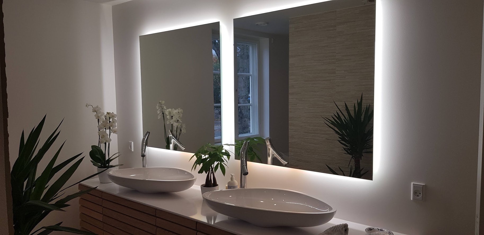Nowoczesna łazienka z dwoma umywalkami nablatowymi o owalnym kształcie, podświetlanymi lustrami i drewnianą szafką pod umywalkami, uzupełniona o rośliny doniczkowe i białe ściany.