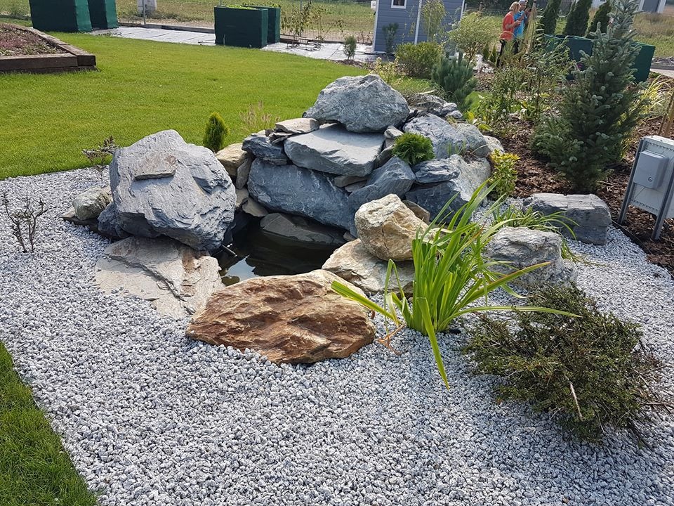 Niewielki, kaskadowy zbiornik wodny w ogrodzie, otoczony szarymi kamieniami i roślinnością, z widocznym trawnikiem w tle.
