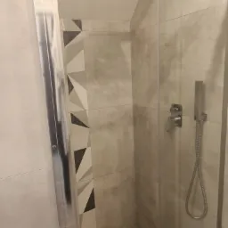 Nowoczesna kabina prysznicowa z geometrycznym wzorem płytek na ścianie, szklane drzwi przesuwne i bateria natryskowa ze słuchawką.